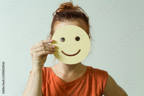 Person holding a yellow smiley face mask, concealing their true emotions.