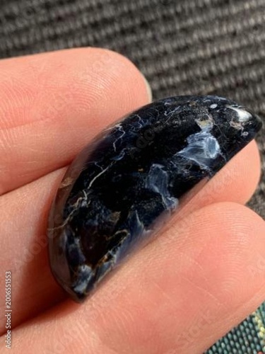 Natural Pietersite Cabochon. A glossy, dark blue stone with white streaks rests in the palm of a hand. The stone is smoothly polished, showcasing its natural pattern.