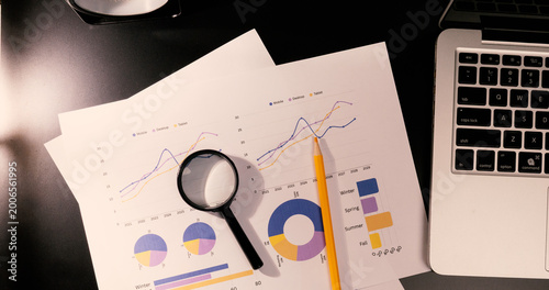 Top view of financial documents with colorful graphs, black magnifying glass, and laptop on dark office desk