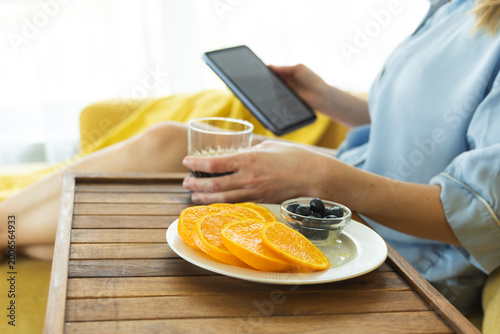 Woman relaxing at home using a tablet, drinking beverage and eating healthy snack with fresh fruit, cozy modern lifestyle and self-care concept