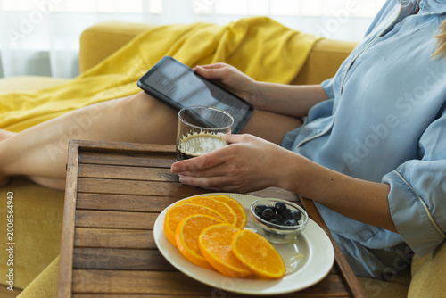 Young woman enjoying healthy breakfast on sofa with coffee and fresh fruits, using tablet at home. Cozy morning routine, self care, relaxation and modern lifestyle concept