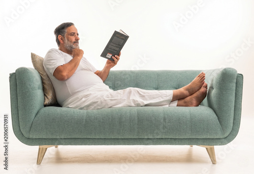 A Saudi man lies on a green sofa reading a book against a plain white background. The subject wears traditional white indoor clothing while resting his head on a beige pillow.
