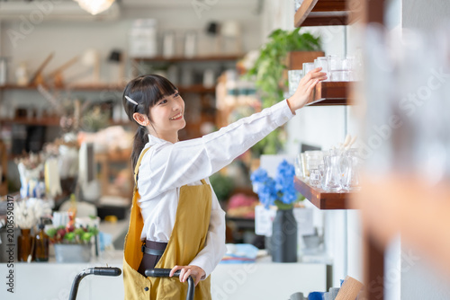Cheerful young Asian woman in a yellow apron browsing and selecting sustainable glass containers on a wooden shelf at a modern zero waste store with a bright atmosphere.