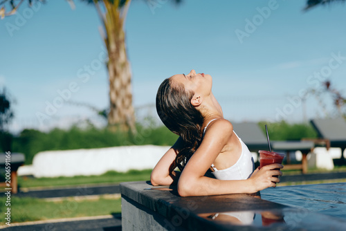 Tableau sur toile Young woman in white swimsuit relaxes by poolside leaning on edge holds cold drink enjoys sunny summer day with eyes closed outdoors in tropical lifestyle setting