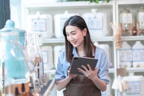 Cheerful young Asian female entrepreneur in a brown apron managing inventory with a digital tablet at a modern boutique store featuring eco-friendly products and refill stations.