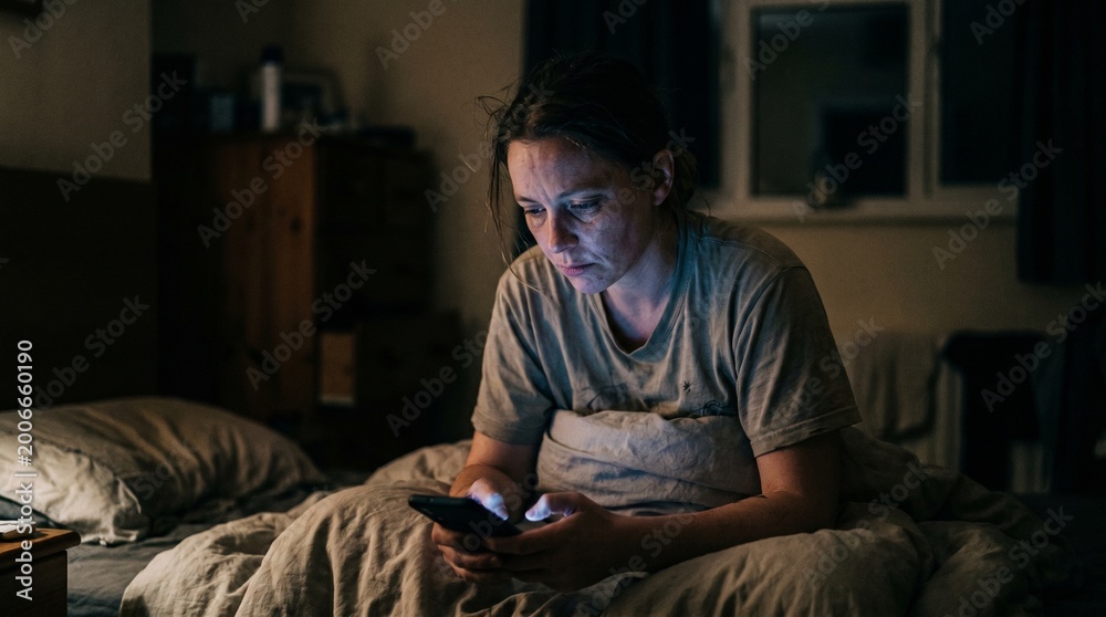 Fototapeta premium A tired young woman with dark circles under eyes using smartphone in bed at night