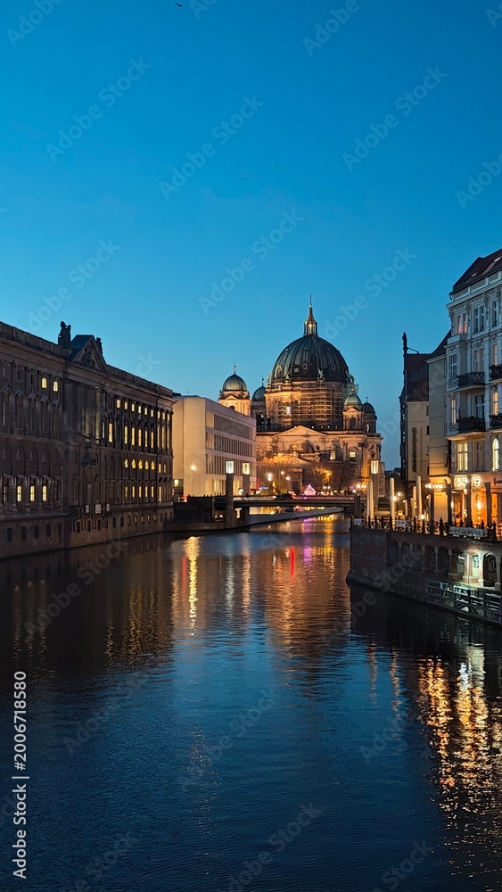 Fototapeta premium Historic buildings line a canal in Berlin during dusk, showcasing illuminated facades and reflections on the water's surface under a clear blue sky