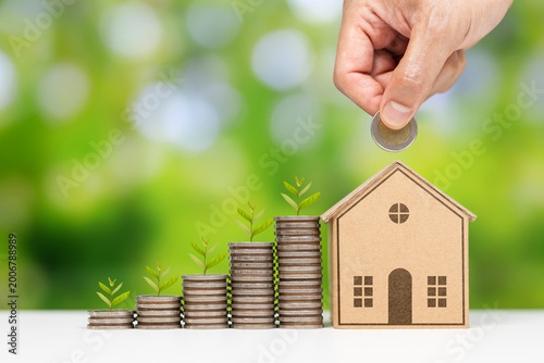 A human hand placing a coin into a small wooden house model positioned in front of rising stacks of coins with small green plants growing on top