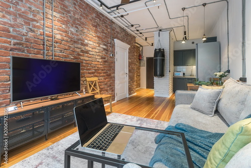 A cozy apartment features a living room with a brick wall and a television. A laptop rests on a table. The kitchen can be seen in the background, showing modern design