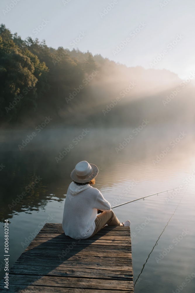 Fototapeta premium Person fishing while sitting on wooden dock at misty lake