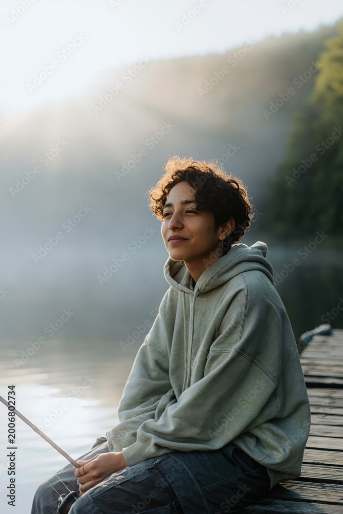 Fototapeta premium Young person sitting relaxed holding fishing rod on dock at misty lake