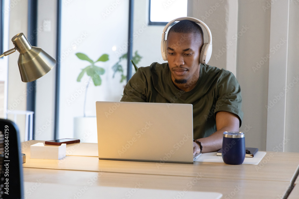 Fototapeta premium African American man typing on silver laptop at wood desk wearing headphones with tumbler nearby