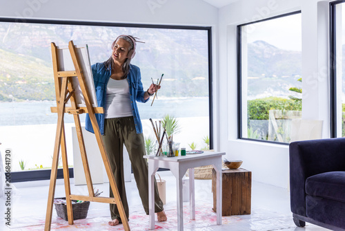Middle-aged female artist standing and painting in home studio with easel, palette, headphones
