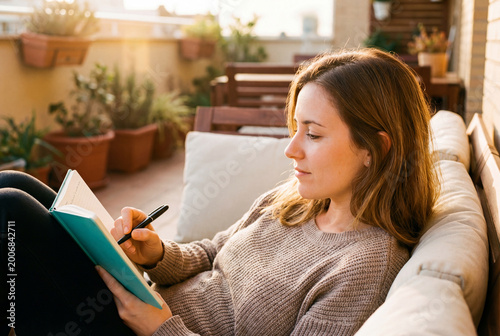 Woman writing in a notebook while relaxing on a cozy terrace at sunset. Concept of mindfulness and reflective lifestyle.