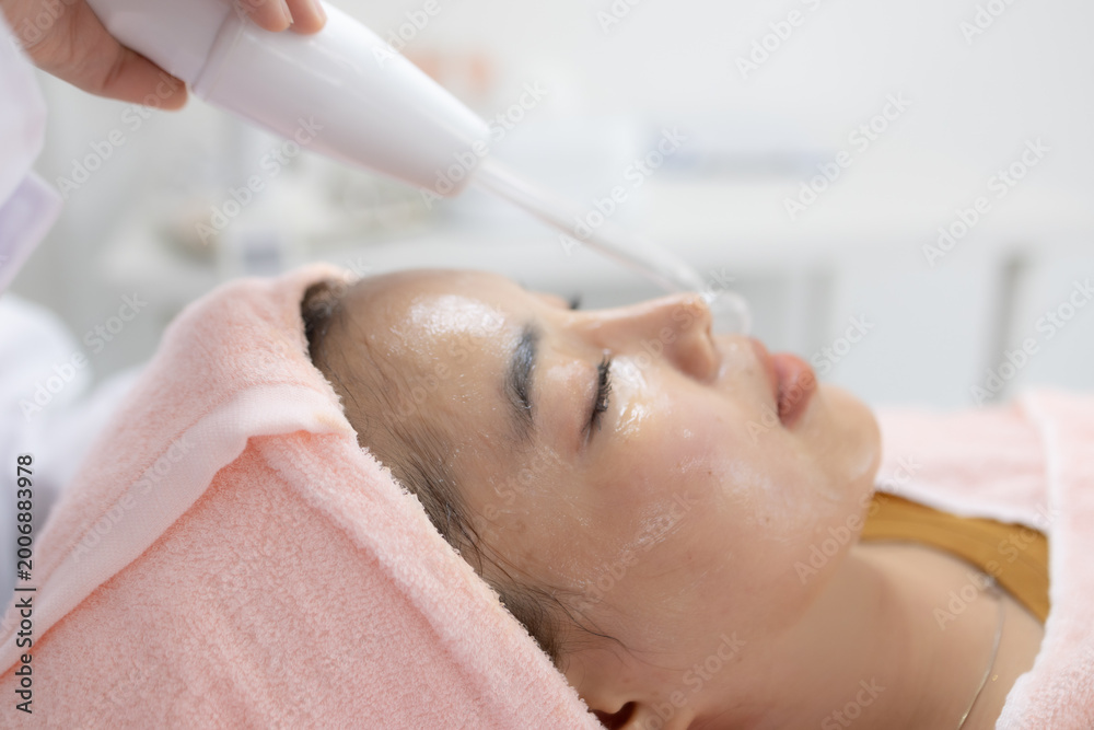 Naklejka premium Soft light glows on calm face. Gentle device delivers soothing mist. Eyes closed, expression serene. Pink towel wraps tenderly around head. Perfect for wellness, spa, or beauty branding 