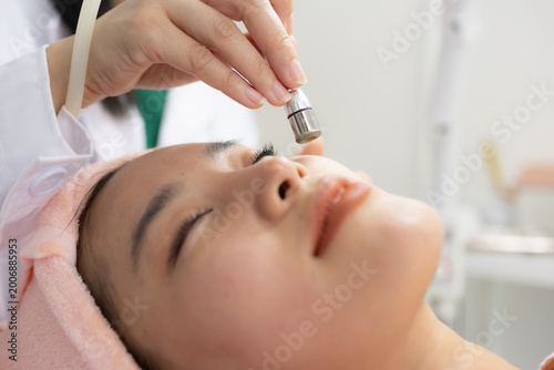 Young woman relaxes during facial treatment. Professional hands use gentle device on her forehead. Her eyes are closed, showing calm and trust. Soft pink towel wraps her head for comfort
