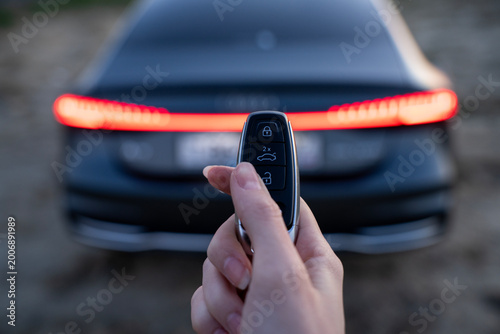 Fotografie Hand pressing trunk button on car key fob near rear of vehicle, with illuminated tail light and blurred automobile background outdoors