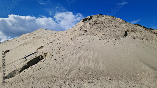 Pile of sand on a construction site under a blue sky. Concept of construction, raw materials and industrial work. Large mountain of gray construction sand against a bright blue sky with light clouds