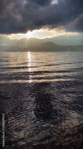 Vertical format of rippling water at sunset, with the sunlight casting a sparkling reflection in the water, with mountain and heavy cloud in the background.