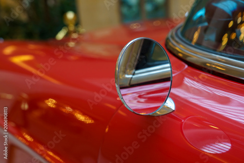 Close-up view of a vintage red car's side mirror reflecting light, showcasing the glossy paint and chrome details in a well-lit indoor setting