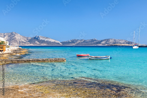 Turquoise bay with small boats, rocky shoreline and rugged cycladic hills in Koufonisi island, Greece, Europe, under a clear blue sky.