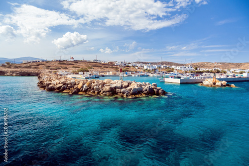 Turquoise harbor waters and rocky breakwater in Koufonisi island, Greece, with fishing boats, yachts, white village houses and a windmill.