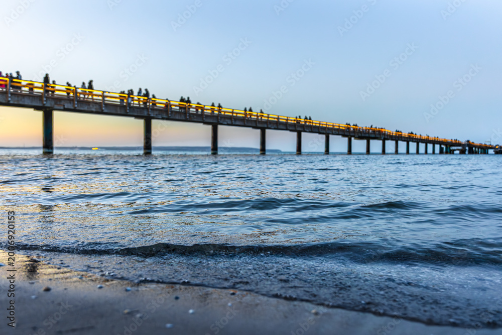 Obraz premium Long sea pier in evening twilight. Waves on foreground during sunset at the beach.