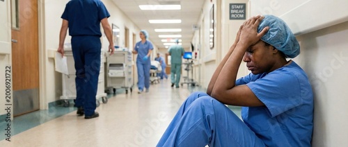 Overwhelmed nurse crying and holding head in hands while sitting on hospital floor, with colleagues walking in hallway