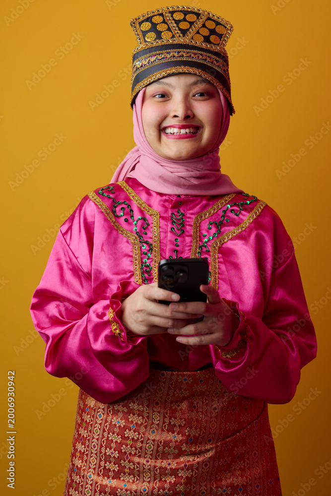 Obraz premium Smiling Young Muslim Indonesian Asian Woman In Bright Traditional Costume And Hijab Holding Smartphone For Celebration