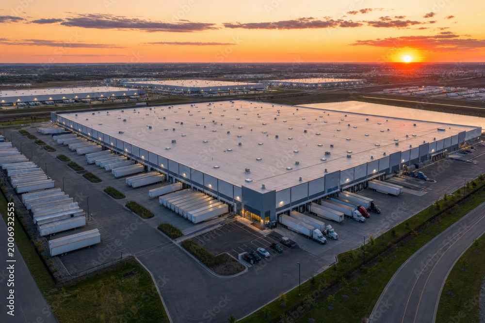 Fototapeta premium Modern distribution center at sunset with numerous trucks at loading docks. High-angle view showcasing busy logistics hub and vast industrial complex operations.