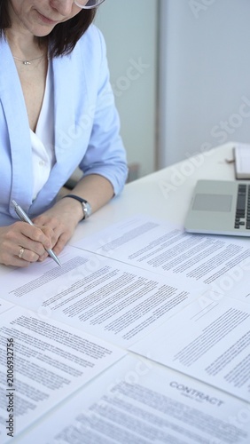 Professional woman in light blue blazer reading and signing important business contract documents, reviewing fine print for agreement and legal compliance at her office desk