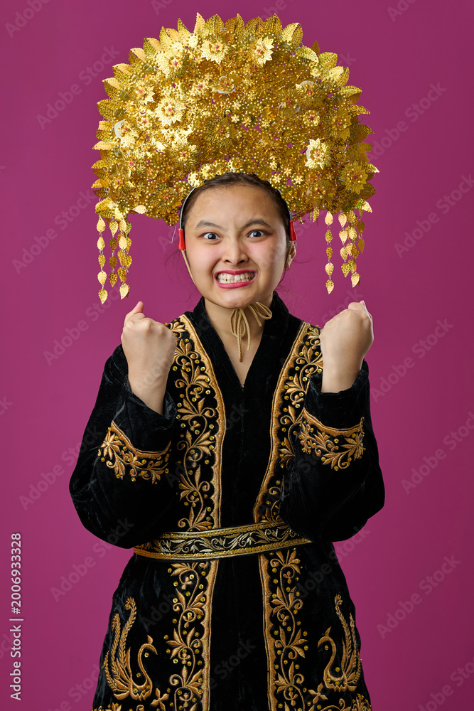 Obraz premium Young Indonesian Asian Girl in Traditional Gold Headdress and Embroidered Robe Celebrating With Excited Expression