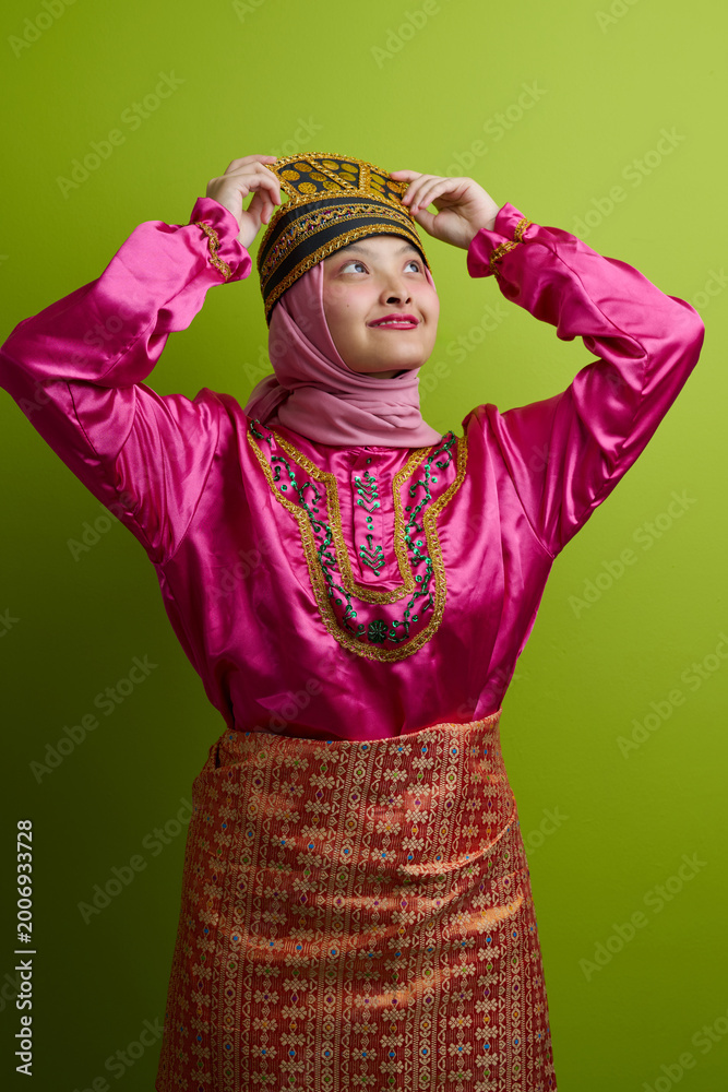 Fototapeta premium Young Indonesian Asian Woman Wearing Traditional Malay Costume With Embroidered Hat And Pink Hijab On Green Background