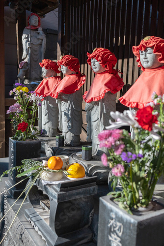 Row of small Jizo statues wearing red bibs and hats with offerings of flowers and fruit at a Japanese temple, capturing traditional Buddhist culture, prayer, and everyday spirituality in soft natural 