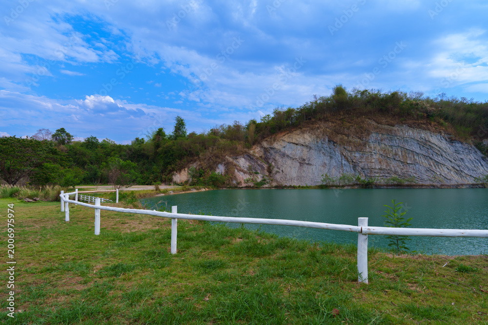 Fototapeta premium Scenic Rural Landscape with White Wooden Fence, Turquoise Pond, and Rocky Cliff under Blue Sky