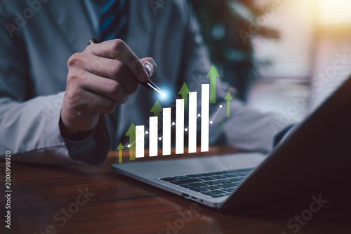 Business growth concept with rising bar chart and upward arrows, hand analyzing financial data on laptop, investment success, profit increase, digital economy and strategic planning visualization.