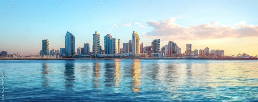 Fototapeta premium Modern coastal city skyline of glass skyscrapers reflected in calm blue water at golden hour with pastel sky and soft clouds, serene mood