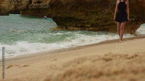 A woman walks barefoot along a sandy beach as ocean waves gently wash ashore. Marinha Beach, Algarve, Portugal