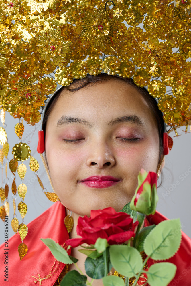Fototapeta premium Young Indonesian Asian Woman Wearing Ornate Gold Traditional Headdress and Red Robe Holding a Red Rose