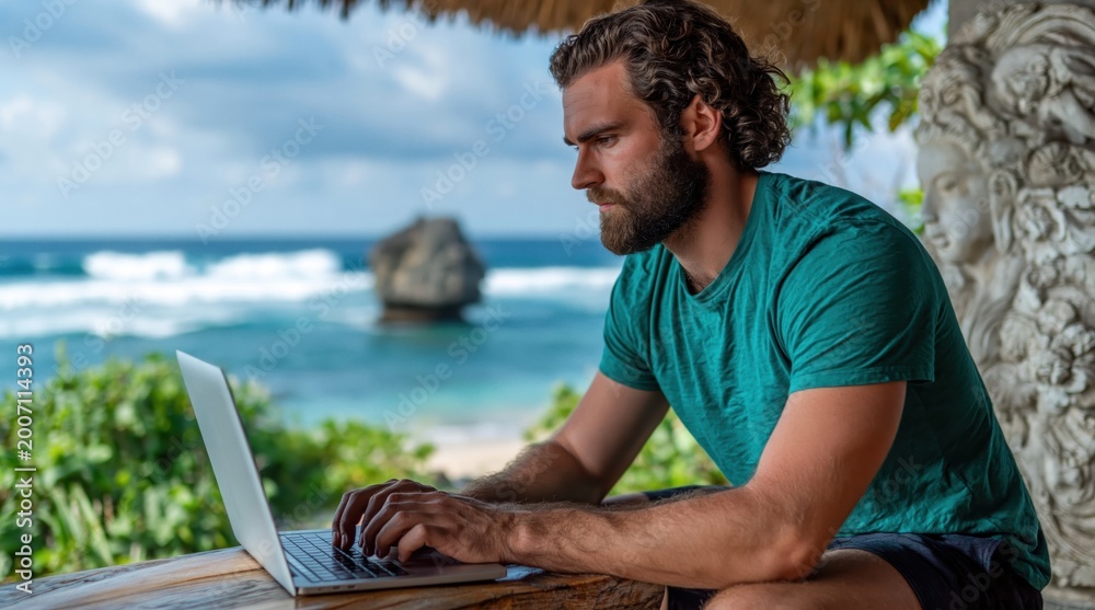 Fototapeta premium Bearded man focused on his laptop, working outdoors with stunning ocean view and lush greenery. High quality photo