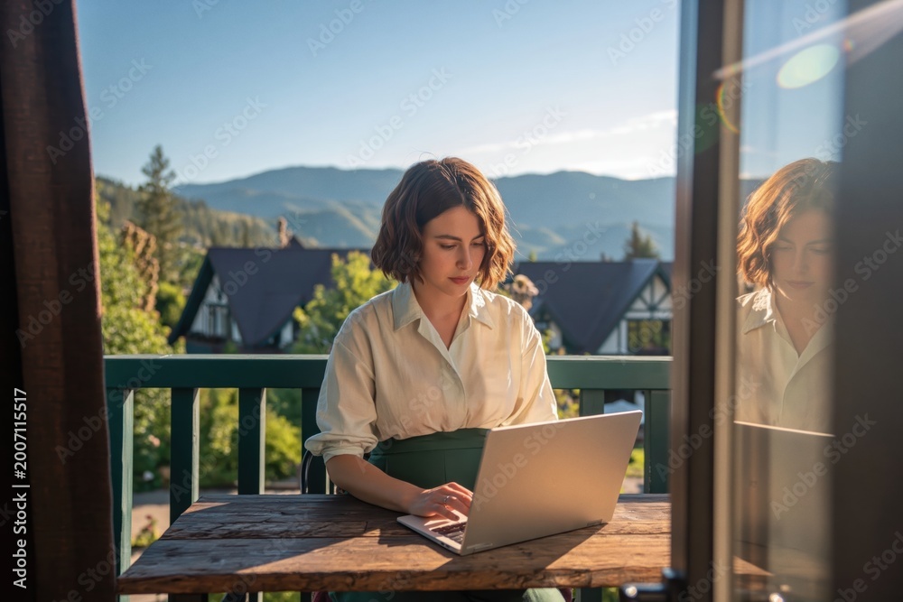 Fototapeta premium Young woman with short brown hair and glasses is focused on her laptop, sitting at wooden table on balcony with stunning mountain view. High quality photo