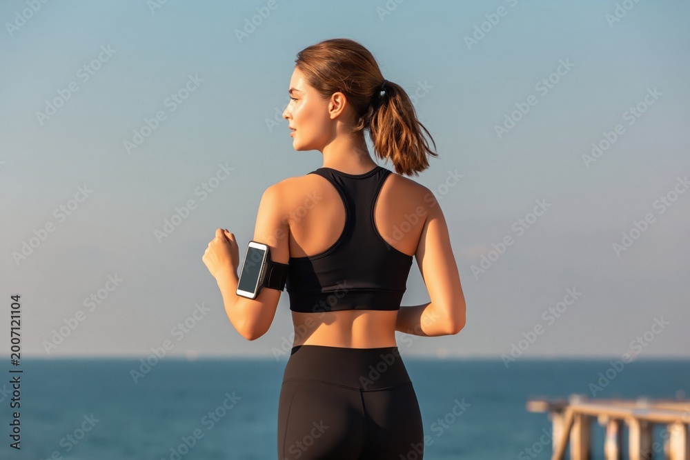 Naklejka premium Young woman exercising outdoors, wearing a sports bra and leggings, running by the sea with a smartphone armband