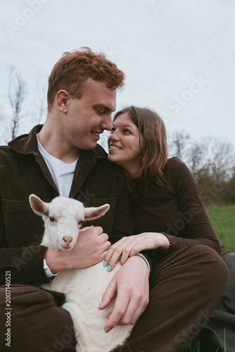 Lifestyle shot of a couple outdoors with a friendly goat kid, smiling and affectionate, sitting in a field, enjoying rural life, warm bond and relaxed moment, natural light, candid vibe, simple