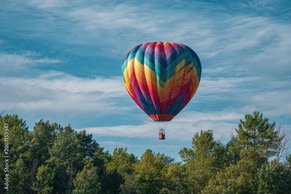 Naklejka premium Vibrant Hot Air Balloon Soaring Above Lush Green Forest Canopy Under a Blue Sky.