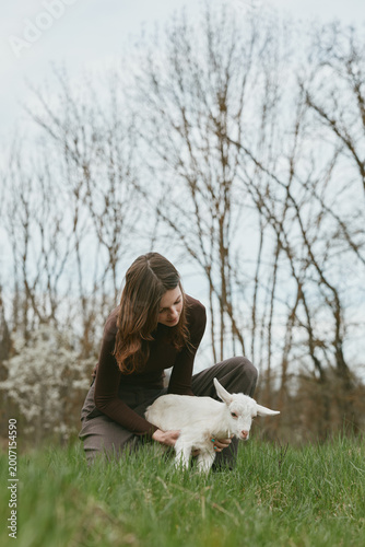 A woman gently pets a white goat kid in a sunlit meadow, capturing a calm countryside lifestyle moment with bond between caregiver and animal reflecting harmony, simplicity, and affection.