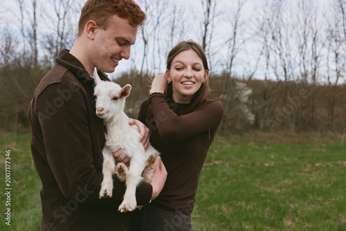 A couple outdoors holds a playful goat kid, sharing a tender moment in a grassy field, capturing lifestyle, happiness, nature, and warm bonding in a rustic countryside scene. Sunny day.