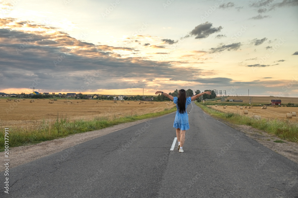 Obraz premium Young woman in blue floral dress stands on dirt road beside golden wheat field at sunset, with distant houses and cloudy sky in the background