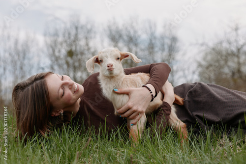 Young woman lying on grass embraces a baby goat outdoors in a natural setting, smiling gently and enjoying a peaceful lifestyle moment in casual clothing.