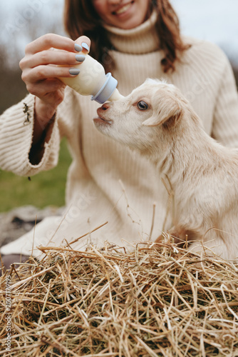 Close-up of a woman feeding a young goat with a bottle, beige sweater, straw background, lifestyle imagery, animal care, maternal bond, warm lighting, farm setting, calm mood, everyday moments