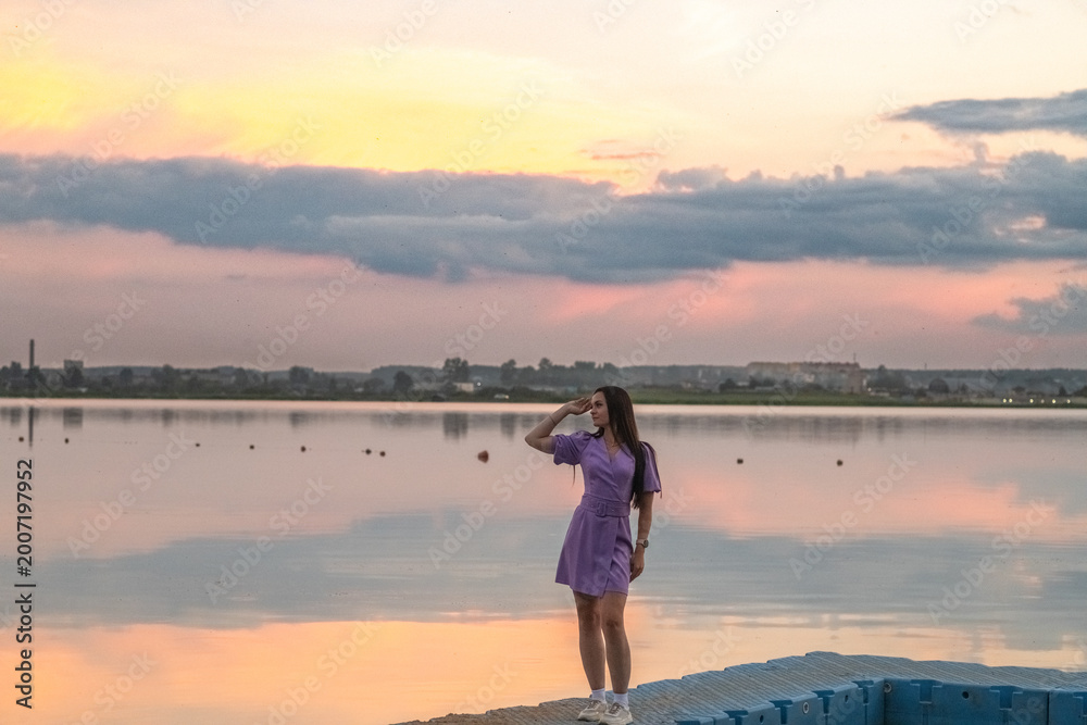 Fototapeta premium Young woman in purple dress stands on blue dock by calm lake at sunset, with dramatic clouds and colorful sky reflecting in the water